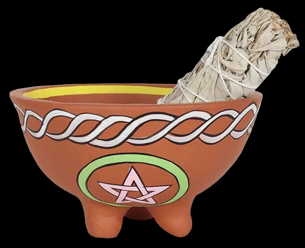 A terracotta-colored clay triple-bowl ritual pot with white Celtic knot patterns, a pink and green pentagram design, and a yellow interior, shown with a white sage smudge bundle inside on a white background.