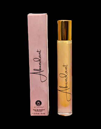 A perfume bottle and its matching pink box are displayed on a wooden surface outdoors, surrounded by white flowers, nuts, and a wicker basket in a blurred garden setting. The bottle has a gold cap and the box is light pink, both featuring the brand name 'Abundant'.