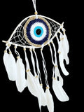 A hanging dreamcatcher with a crocheted evil eye motif in navy, white, and turquoise, surrounded by white feathers and small wooden beads on a light neutral background.