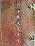 A square brown leather-bound book or journal with embossed circular mandala patterns, featuring a vertical row of colorful gemstone accents down the center and brass corner hardware.